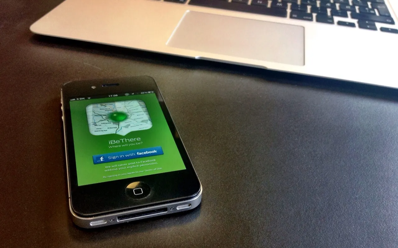 An iPhone 4 displaying iBeThere beside a MacBook on a desk.